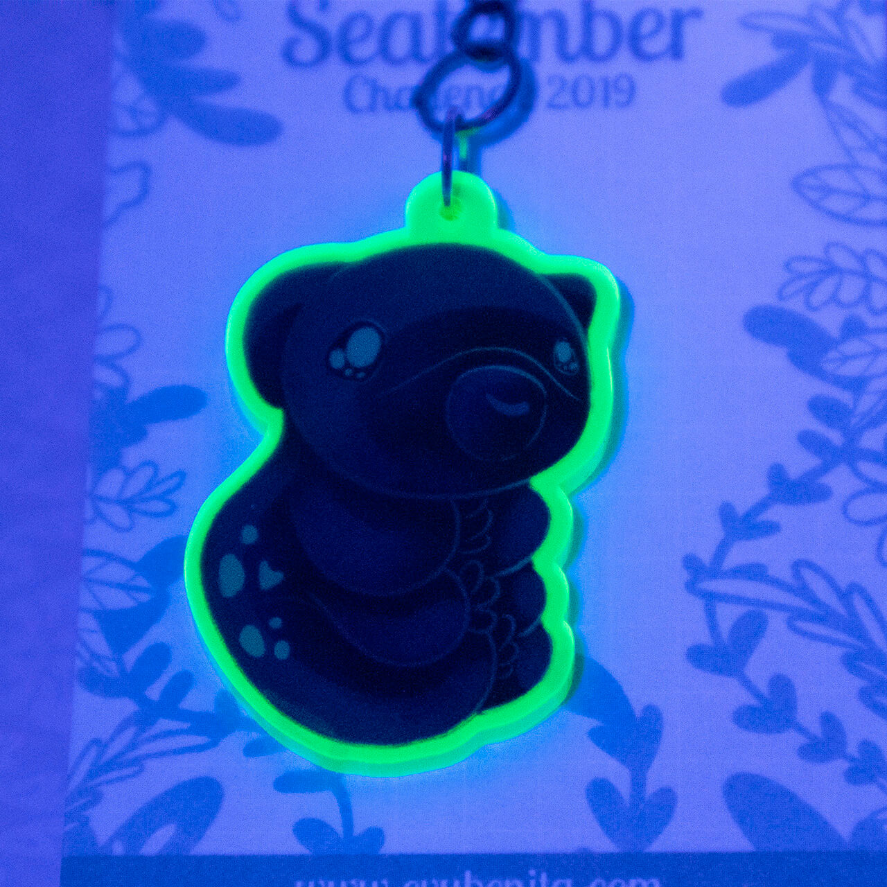 Shows an acrylic charm featuring a cartoon style tardigrade, or "water bear". The charm is made from a material that lights up under UV lamps. The photo demonstrates the vibrant glow.