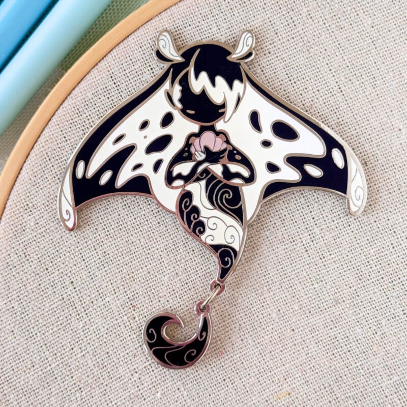 A mermaid enamel pin with a dangle-charm dorsal fin. The mermaid is inspired by the Giant Oceanic Manta Ray. Designed by Evy Benita.