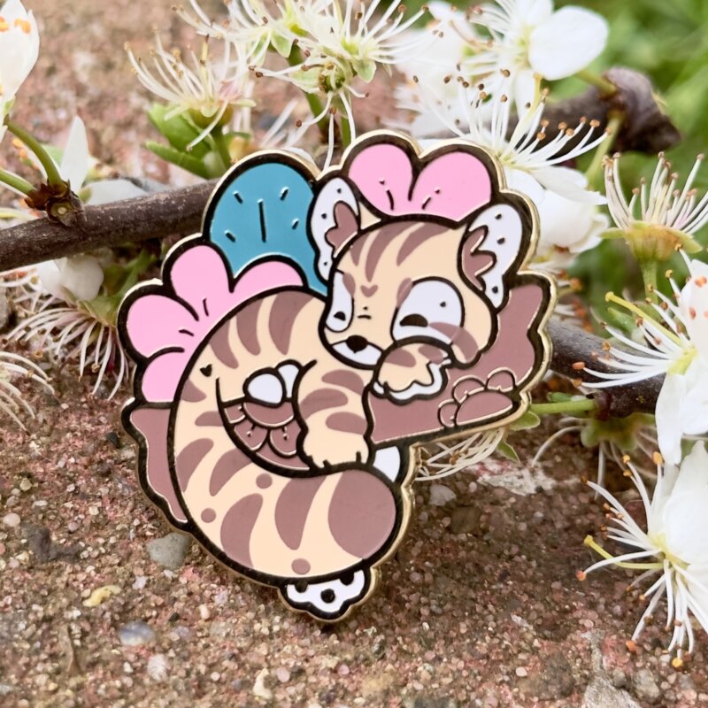 A cute enamel pin showing a cartoon-style sand cat sleeping on a branch. The cat is backed by large pink flowers and a big leaf. Below the cat's tail, you can see toe beans poking through.